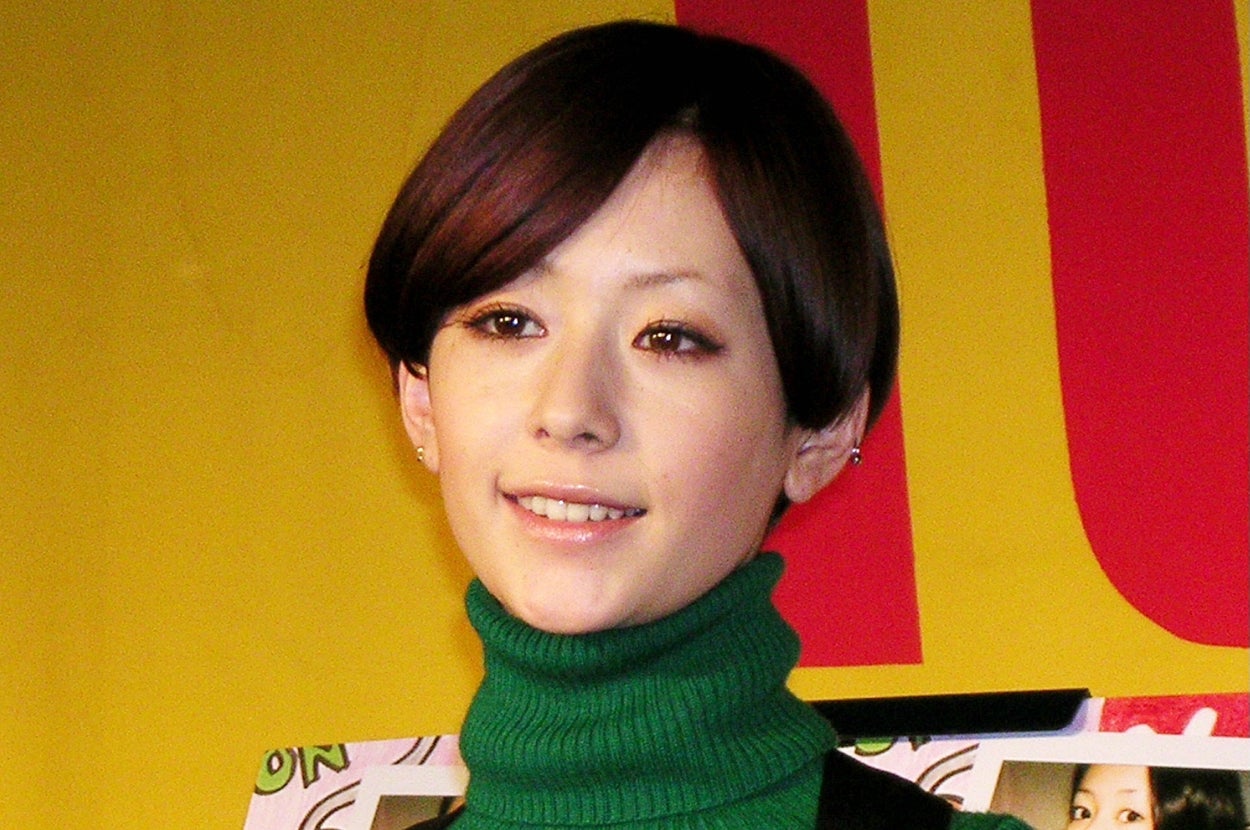 日本の女性、緑のタートルネックに黒の服を着て、写真入りの本を持って微笑んでいる。