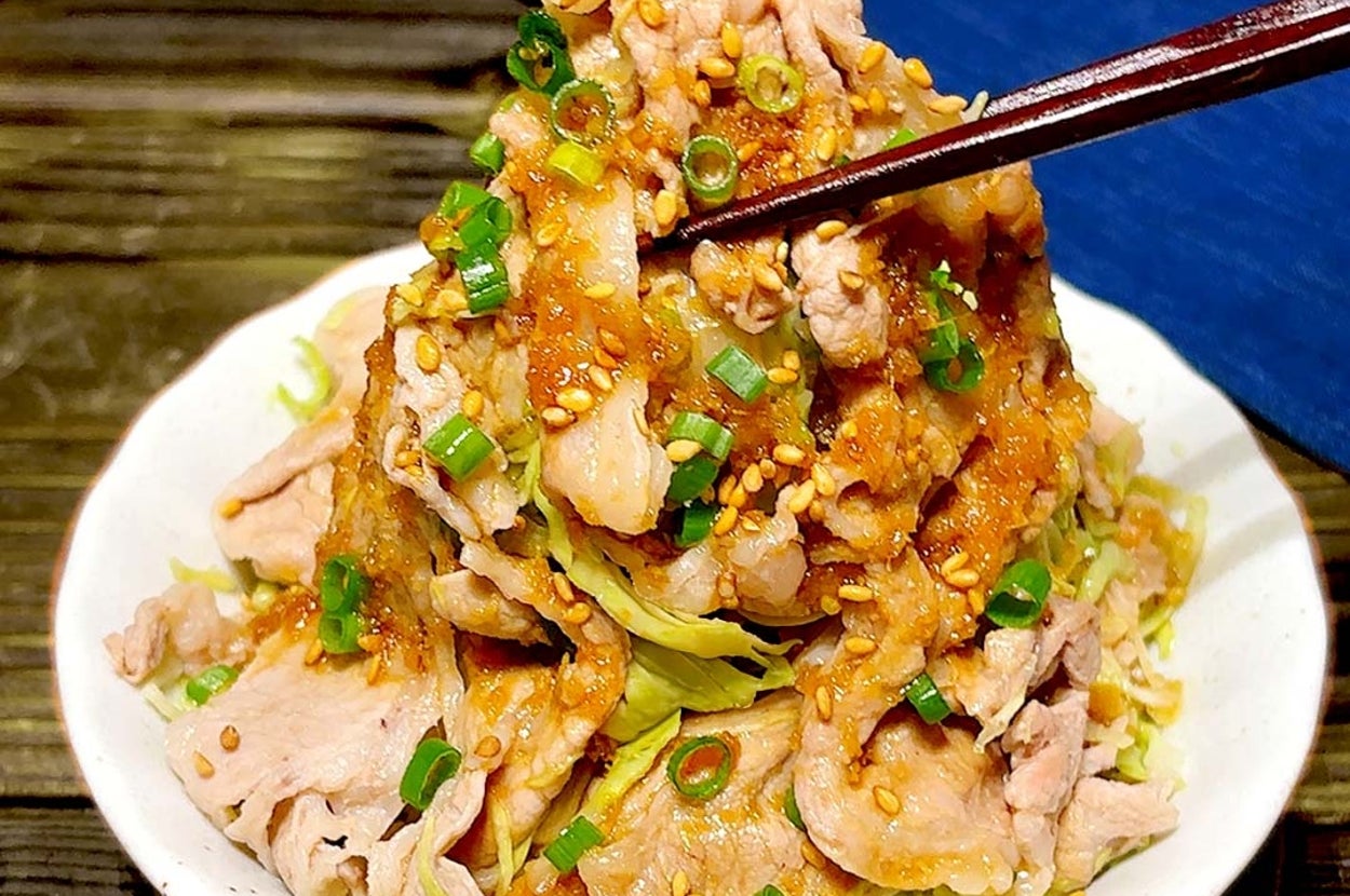 レタスの上に豚肉スライスとネギが盛られ、ゴマとソースがかけられた料理が箸で掴まれている。