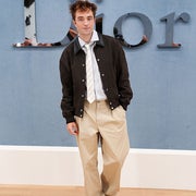 A person in a casual yet stylish outfit with a jacket, tie, and loose pants poses in front of a "Dior" backdrop at an event
