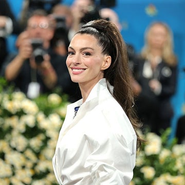 Person on a red carpet smiling, wearing a sleek, white, long-sleeve dress. Background includes photographers and floral decor