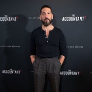 A person stands on a red carpet at a movie event, wearing a fitted long-sleeve shirt and high-waisted trousers