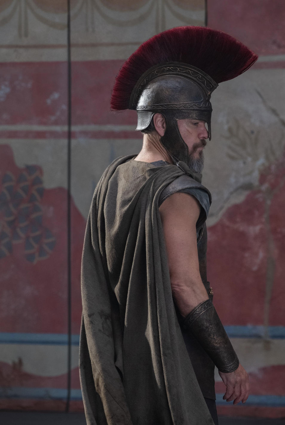 A person in ancient warrior attire with a plumed helmet stands in profile, wearing a draped cloak and metal armor in a historical setting