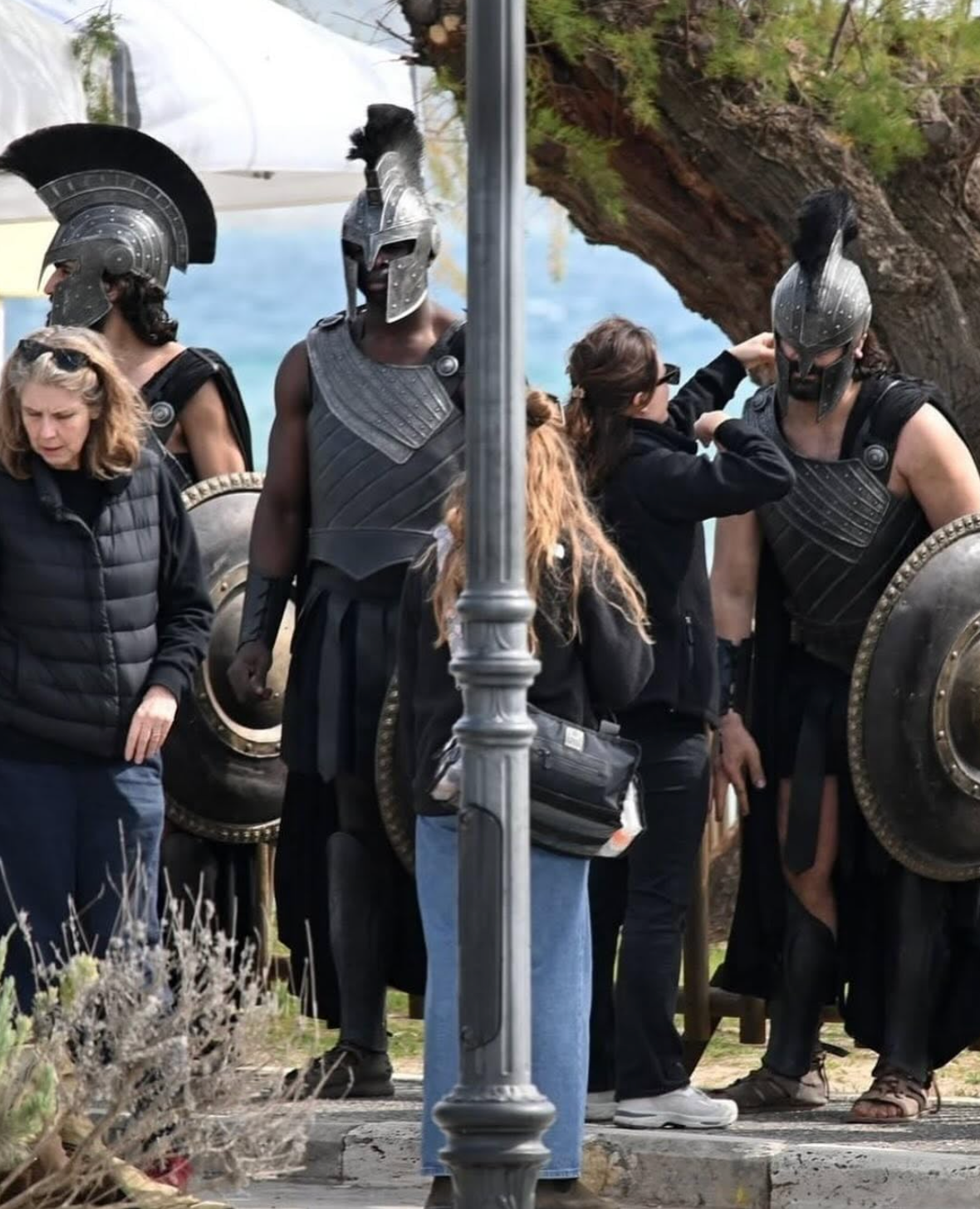 People in ancient warrior costumes stand near a tree, adjusting their armor and helmets. Crew members assist with preparation