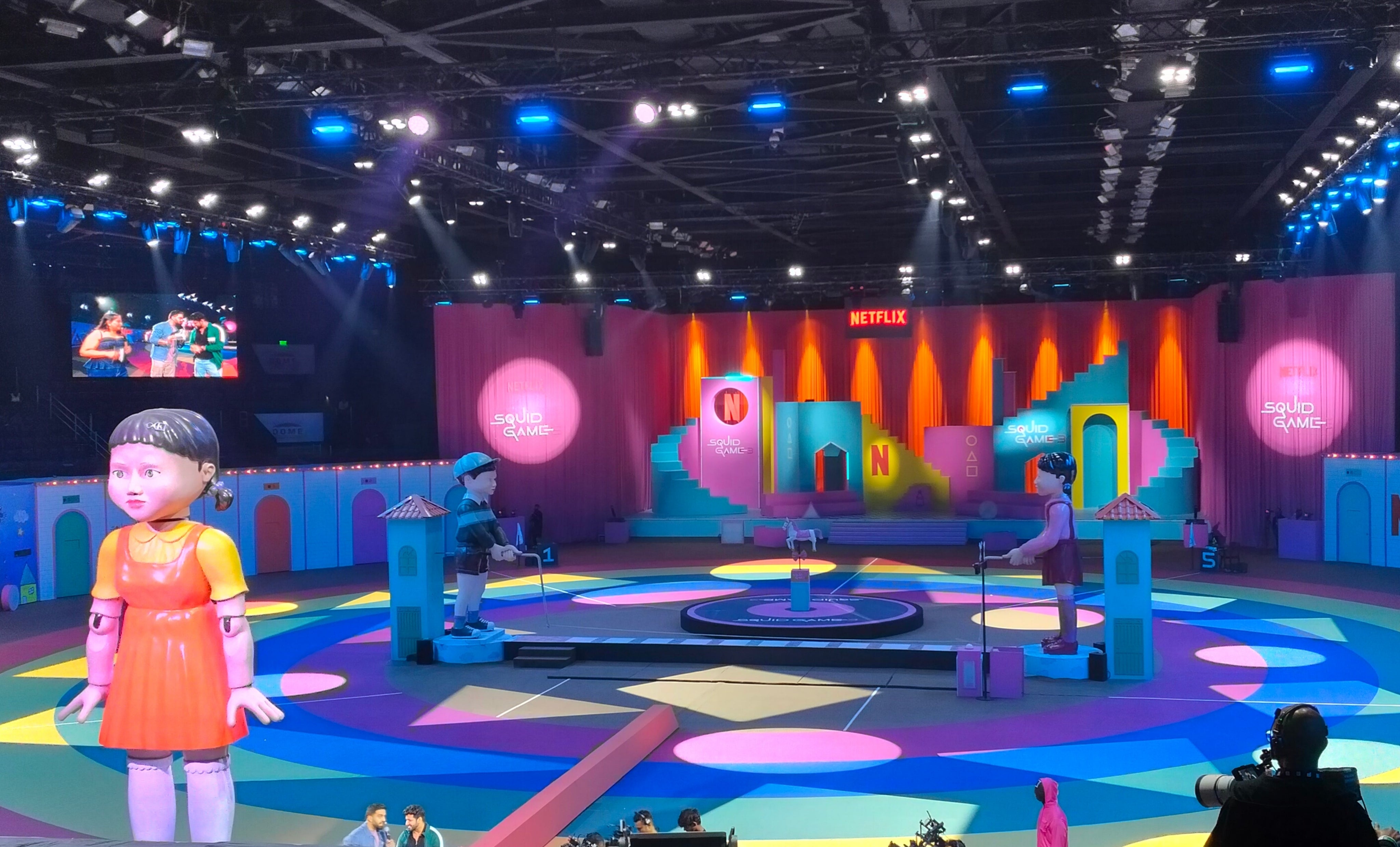 Large event stage with life-size "Squid Game" doll and colorful set; audience seating visible. Vibrant, immersive atmosphere