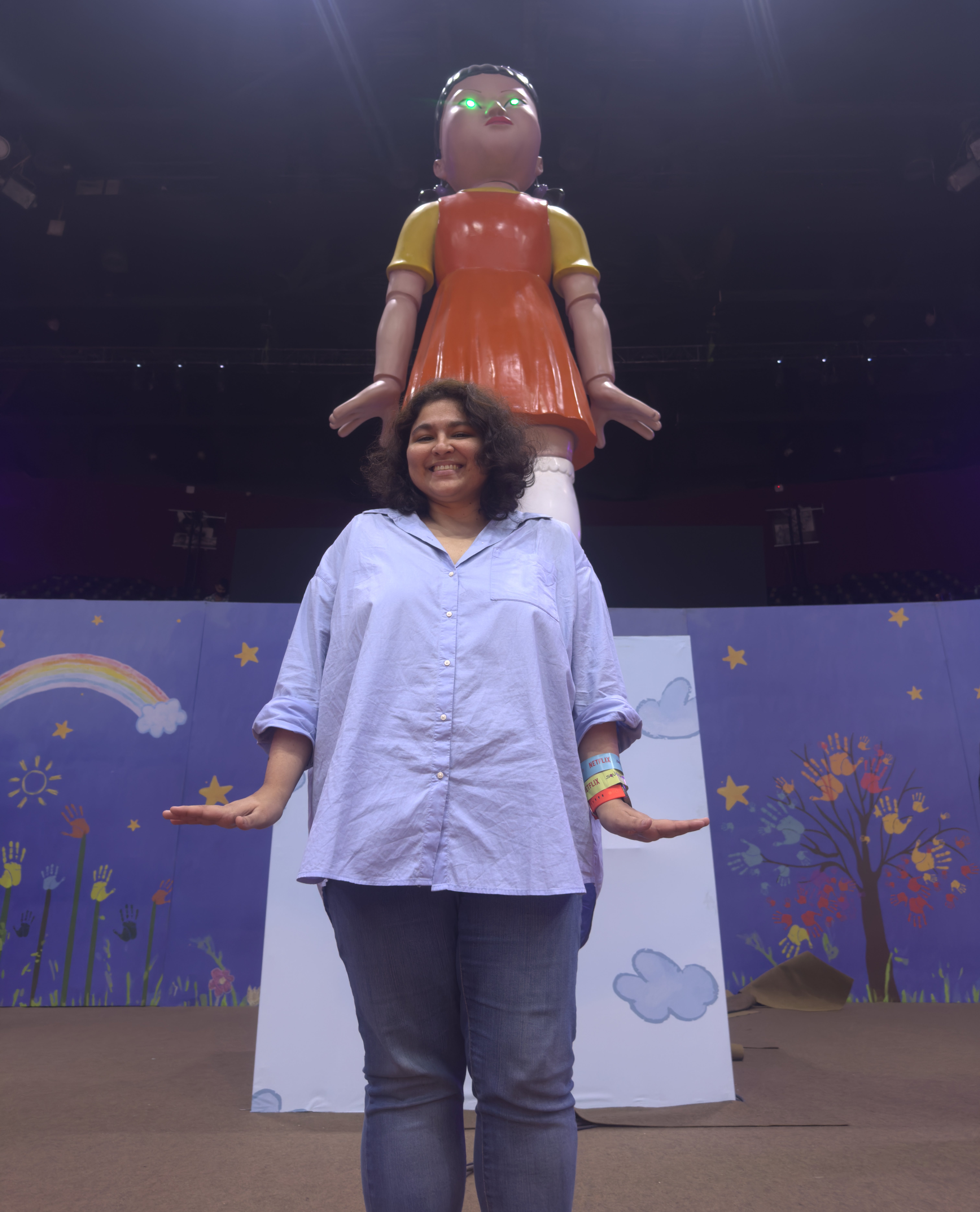 Woman posing in front of large child-like statue with arms raised playfully, indoors with colorful wall decor