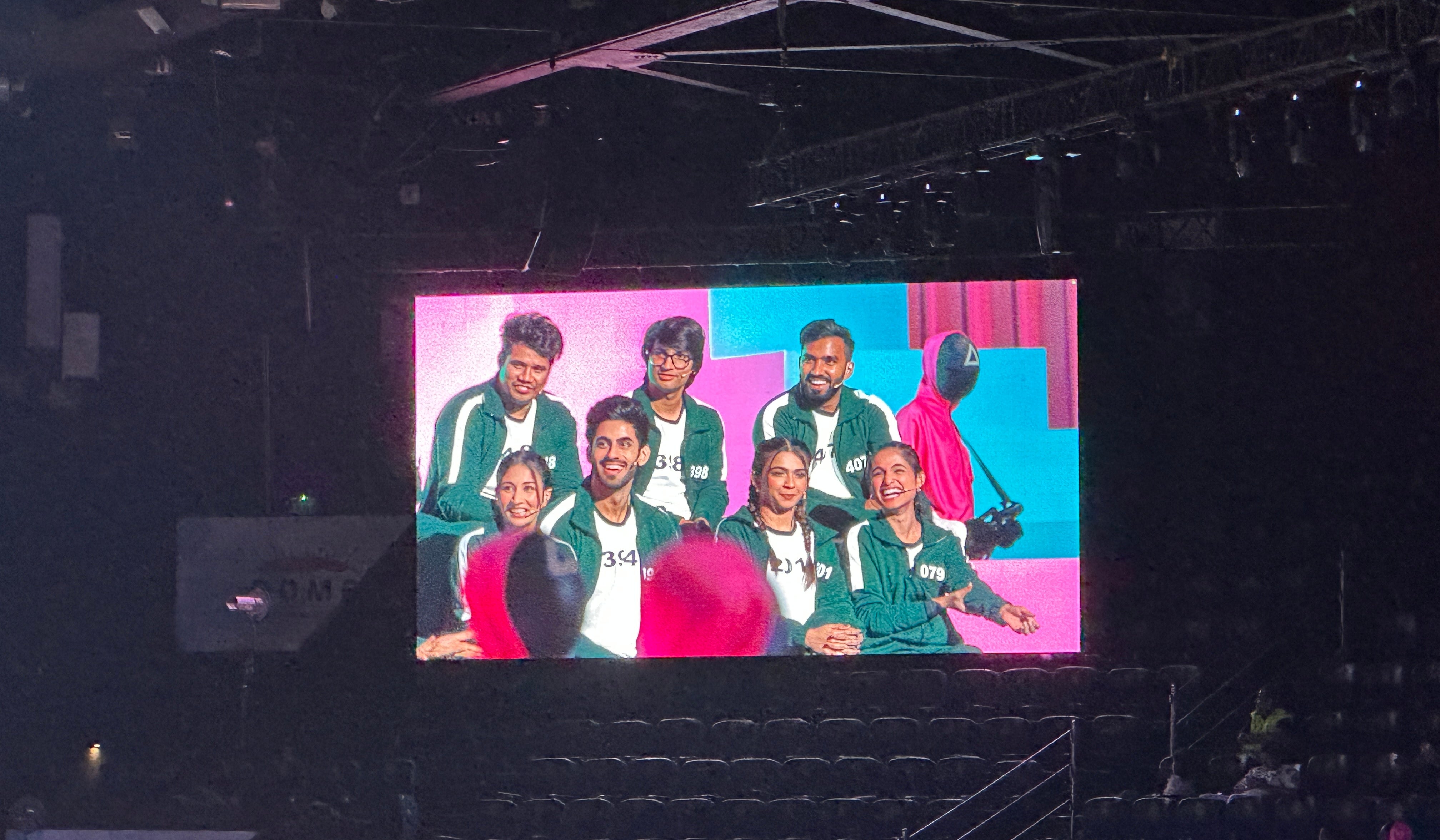 A group dressed in themed tracksuits poses on a large screen at an event, resembling a scene from "Squid Game."
