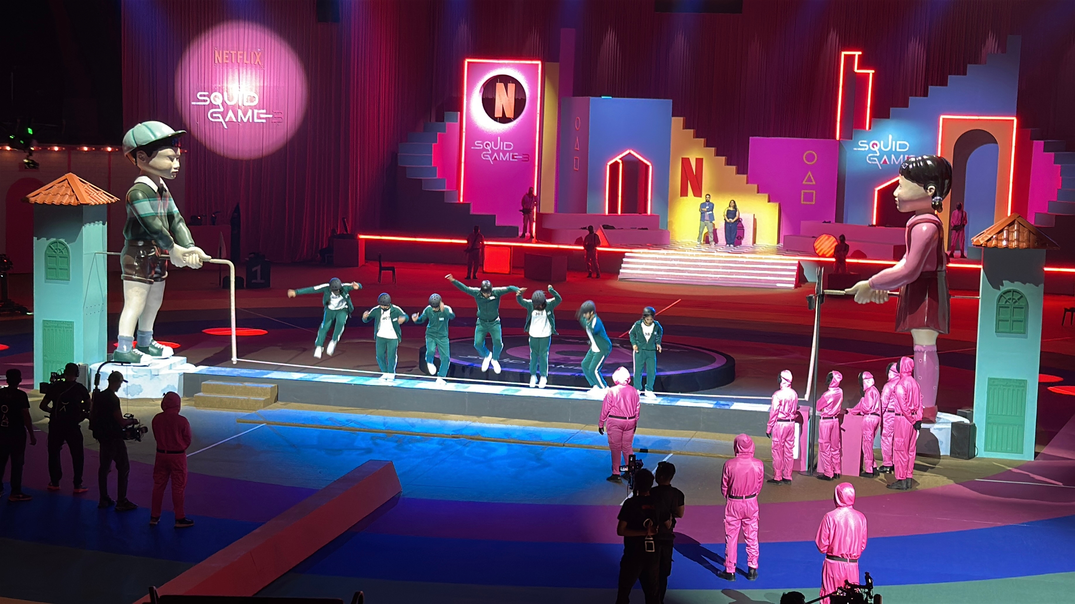Indoor set inspired by "Squid Game," featuring large doll figures and people dressed in series-themed costumes on a colorful stage