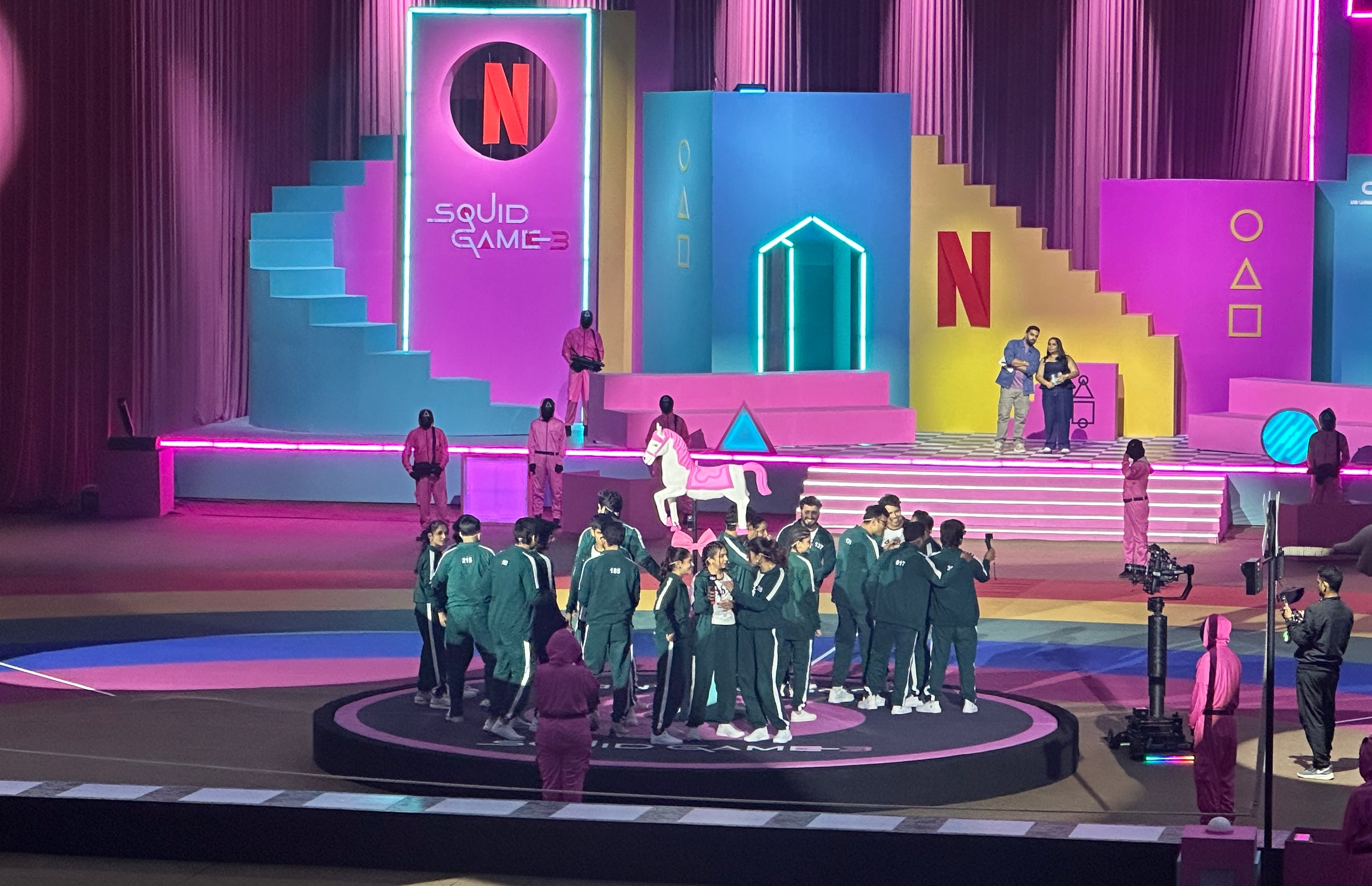 People gather on a stage with "Squid Game" signage. The set has vibrant geometric shapes. Some wear tracksuits; others are in jumpsuits