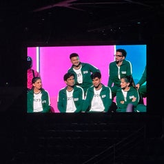 Several individuals on a stage wear matching green athletic jackets, seated and standing, engaged in conversation