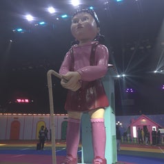 Giant sculpture of a doll in pink clothing holding a cord on a stage with colorful lights and structures in the background