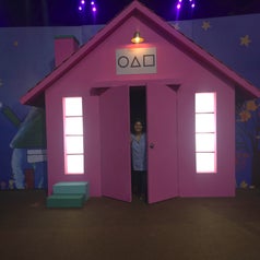 Person standing in the doorway of a whimsical, cartoon-like pink house with geometric shapes above the door