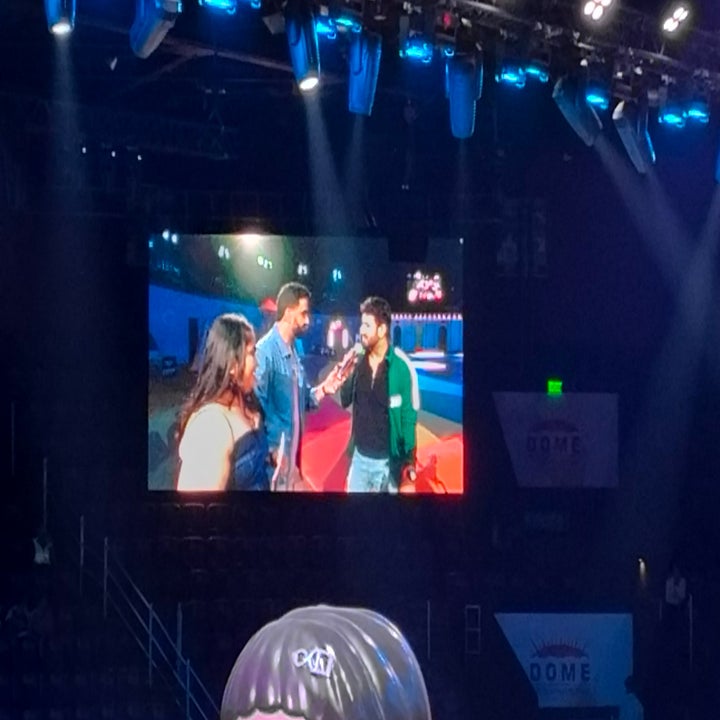 People talking on a stage with a large screen displaying them. The event appears to be in a large venue with bright stage lighting above