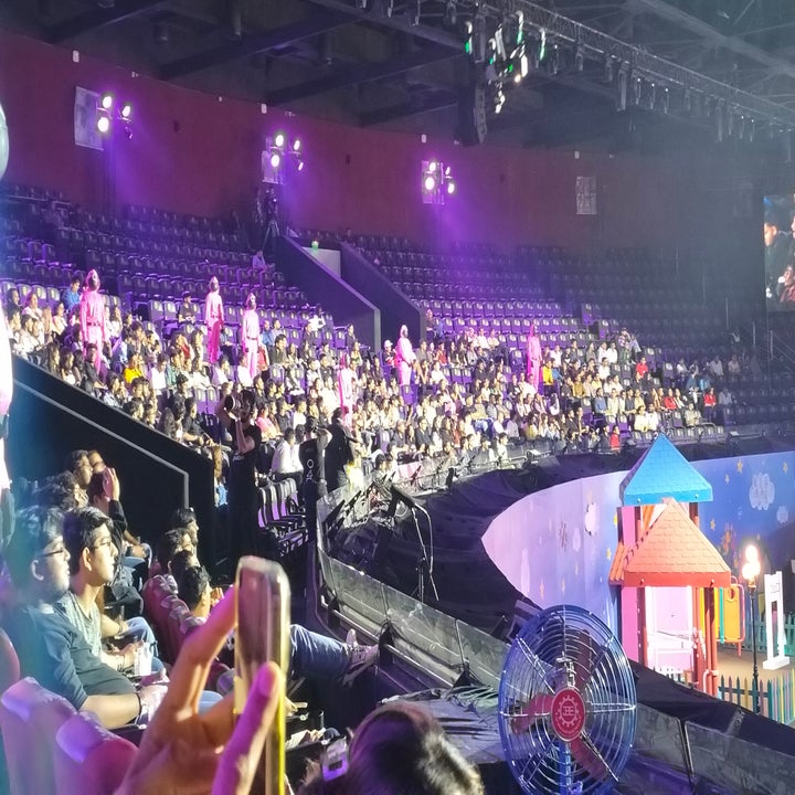 Audience seated in an indoor arena, watching a live event on stage. People are focused on performances with stage lights illuminating the scene