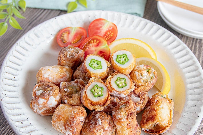 ちくわの中にオクラが入った揚げとトマト、オクラ、レモンが白い皿に盛られた料理。背景に箸と皿。