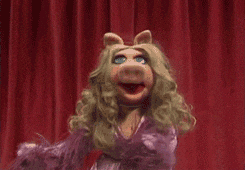 Miss Piggy, the Muppet character, is animated with expressive gestures on a stage with red curtains