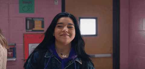Person smiling and walking confidently down a school hallway, wearing a jean jacket and hoodie. Disney+ logo is visible