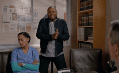 A person in a blue scrubs-looking outfit appears frustrated, sitting next to a joyful individual clapping in an office setting