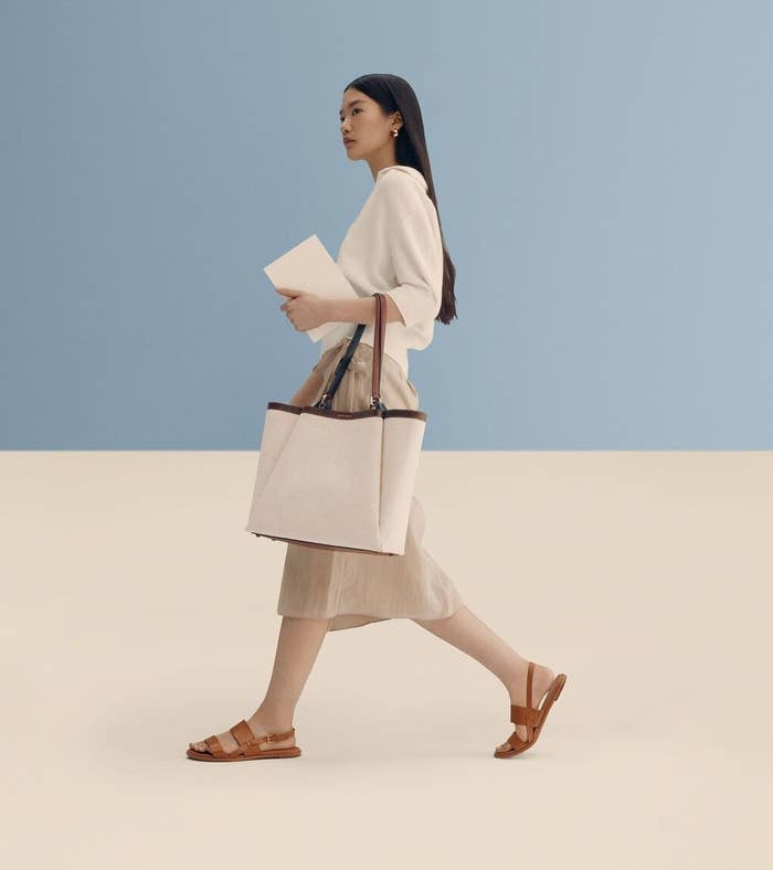 Person walking stylishly with tote bag, wearing a minimalist outfit and sandals, holding a notebook against a elemental background