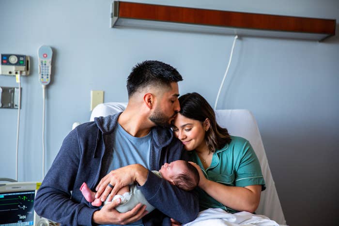 A mates is sitting successful a infirmary room, with the antheral holding and kissing a newborn baby, portion the pistillate lovingly looks astatine the baby