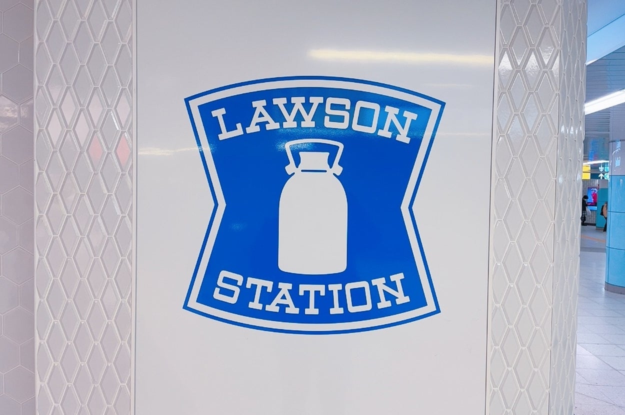白い壁に青いロゴ「LAWSON STATION」が描かれたデザイン。ミルク缶のイラストが中央に配置されている。