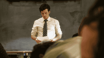 Man in shirt and tie stands in front of a chalkboard, looking at a paper; students sit at desks in a classroom setting