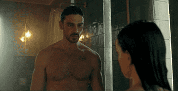 A man stands shirtless in a dimly lit shower scene, facing a woman with wet hair