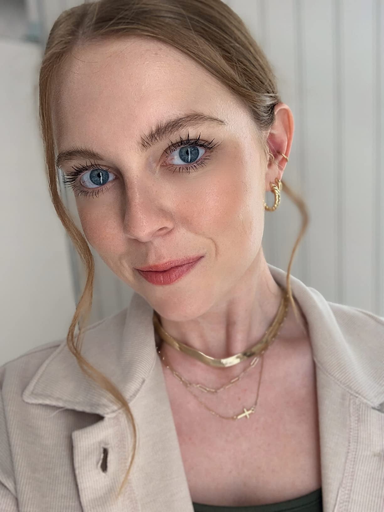 Person with airy  hairsbreadth  tied back, wearing golden  hoop earrings, layered necklaces, and a beige jacket, poses for a close-up photograph  with a brushed  smile