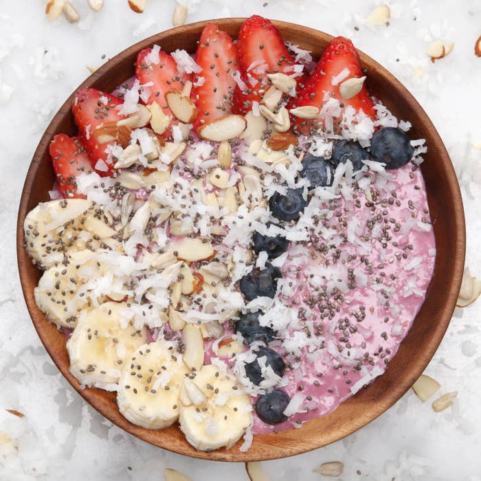 A wooden bowl with a smoothie topped with sliced strawberries, blueberries, banana, coconut flakes, chia seeds, and almond slivers