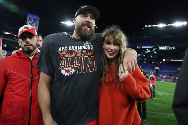 A man in a Chiefs shirt with a cap stands on a field, arm around a smiling woman in a casual knit sweater, after a sports event