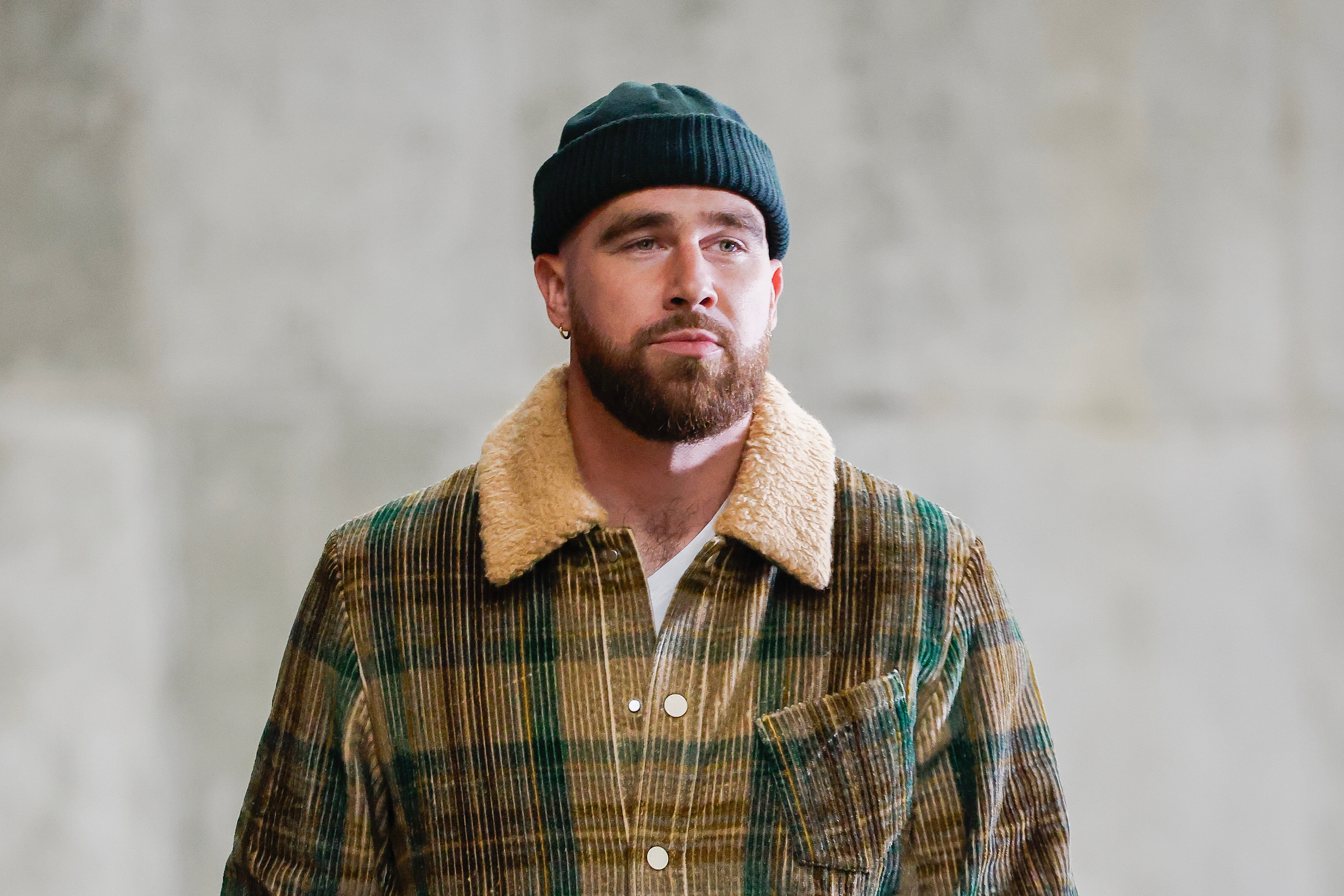 Travis Kelce wearing a plaid jacket with a shearling collar and a beanie stands against a neutral background