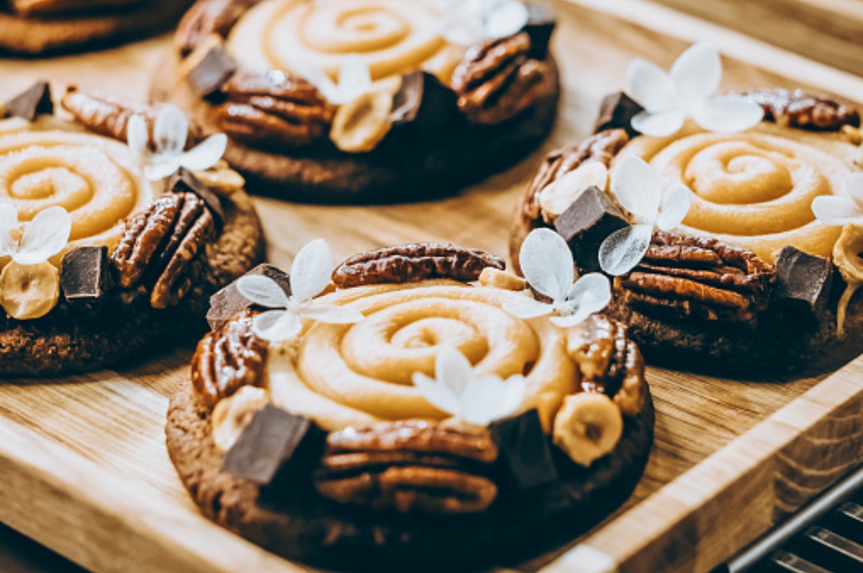 Cookies with caramel swirls, pecans, chocolate chunks, and decorative white leaves on a wooden tray