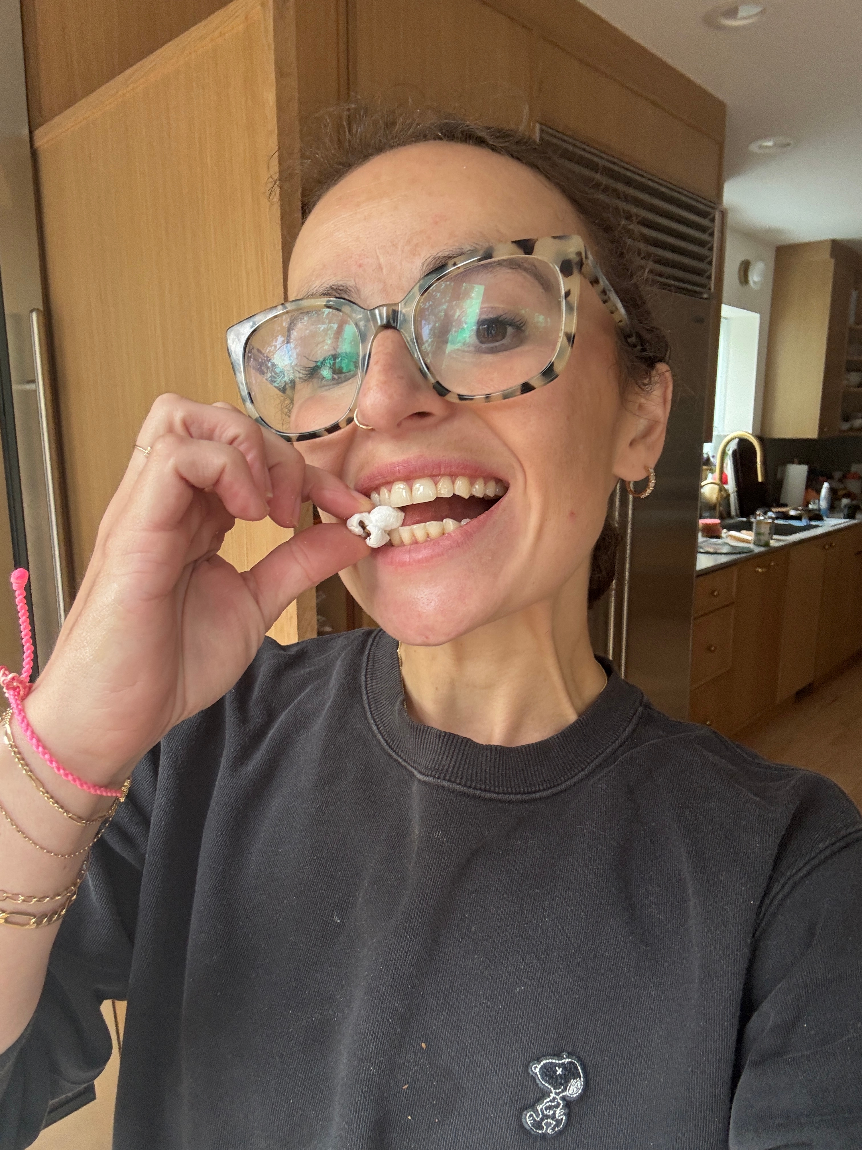 A person with large glasses enjoys a piece of popcorn in a modern kitchen, smiling and wearing a Snoopy sweatshirt