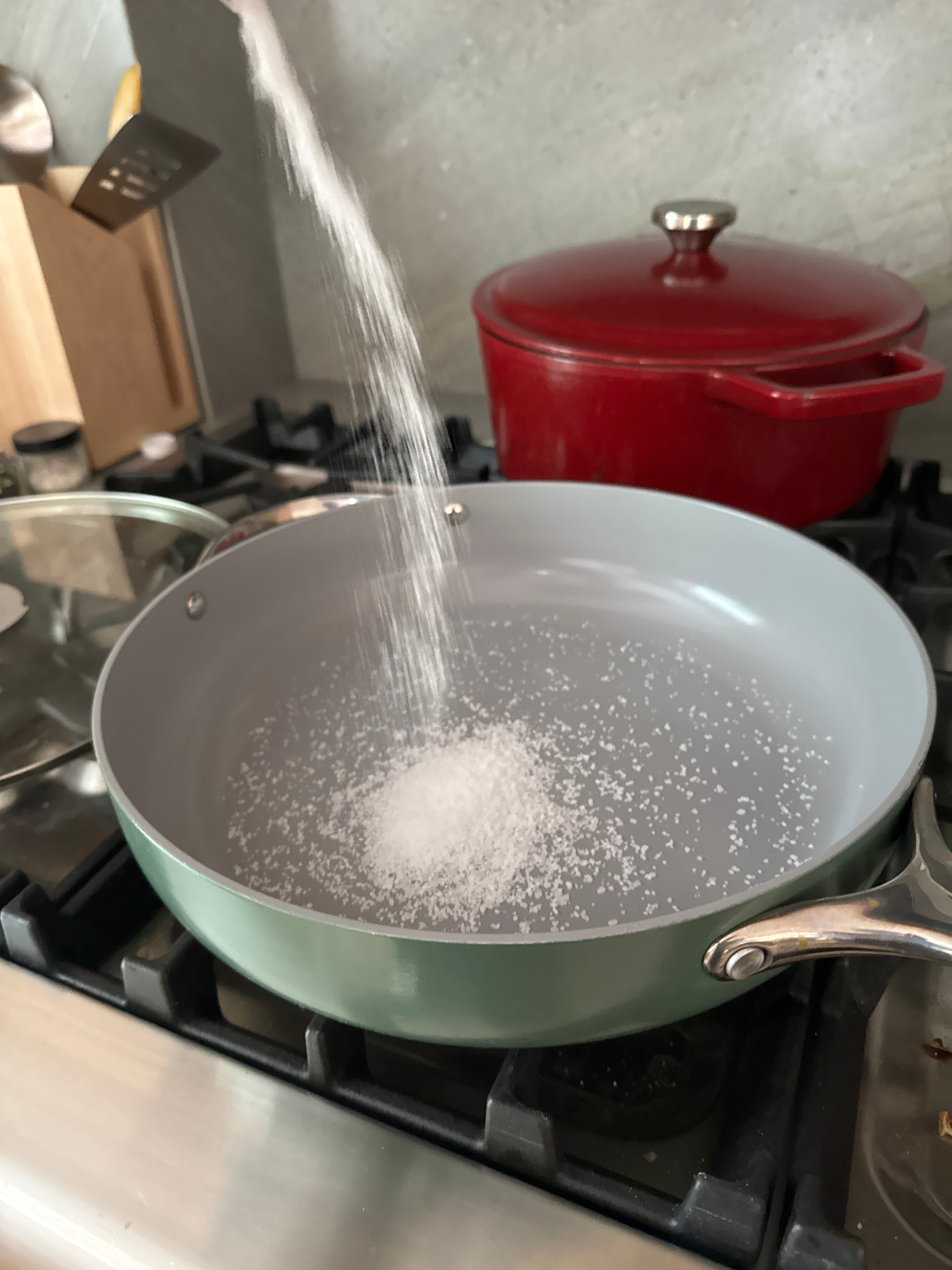 Salt being poured into a frying pan on a stove, next to a red pot and kitchen utensils in the background