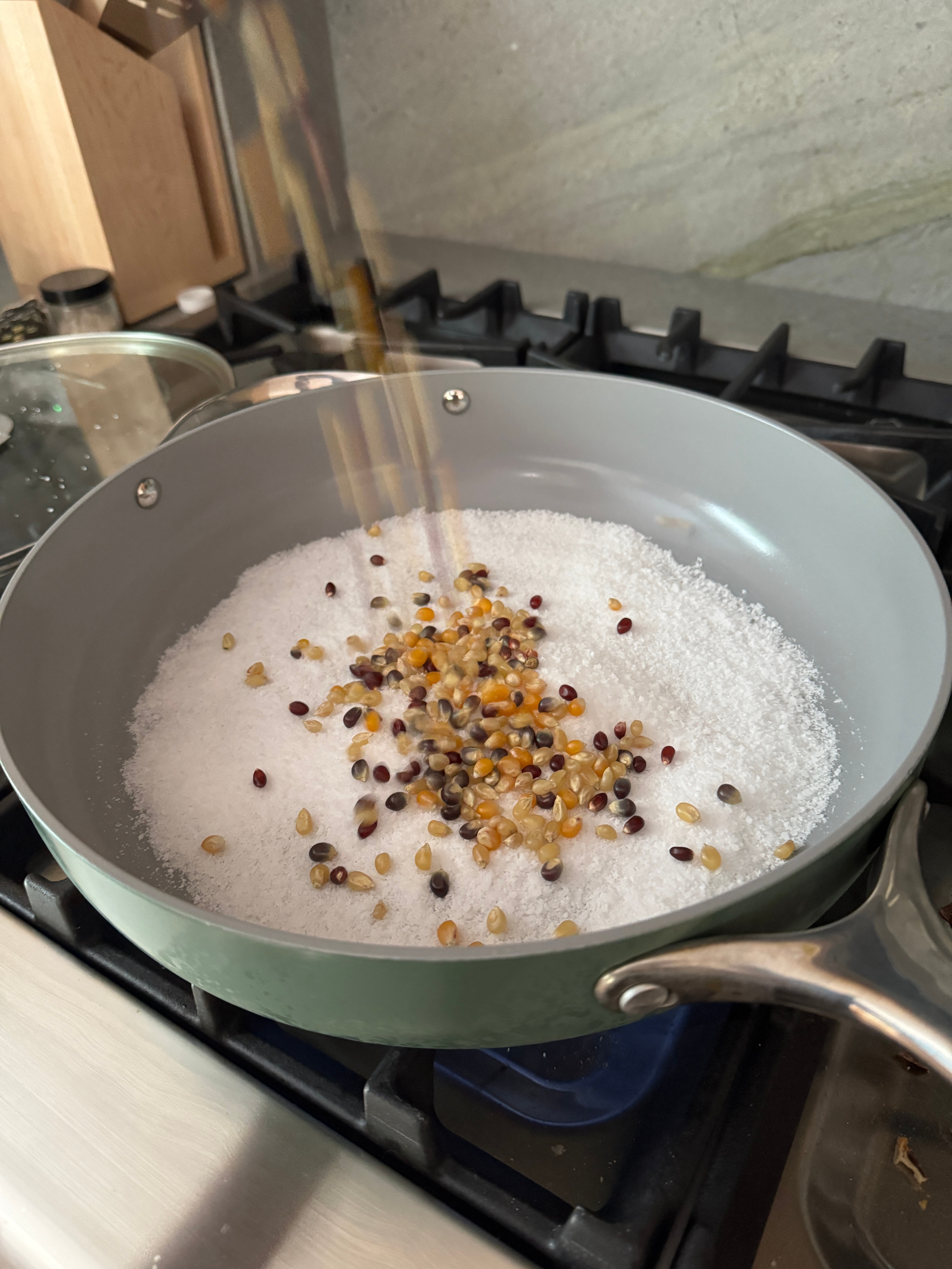 Popcorn kernels falling into a frying pan filled with salt on a stovetop, preparing for popping