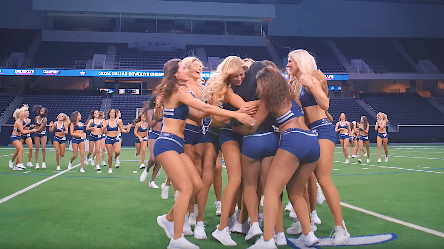 A group of cheerleaders in matching outfits excitedly huddle on a football field, with others visible in the background at an indoor stadium
