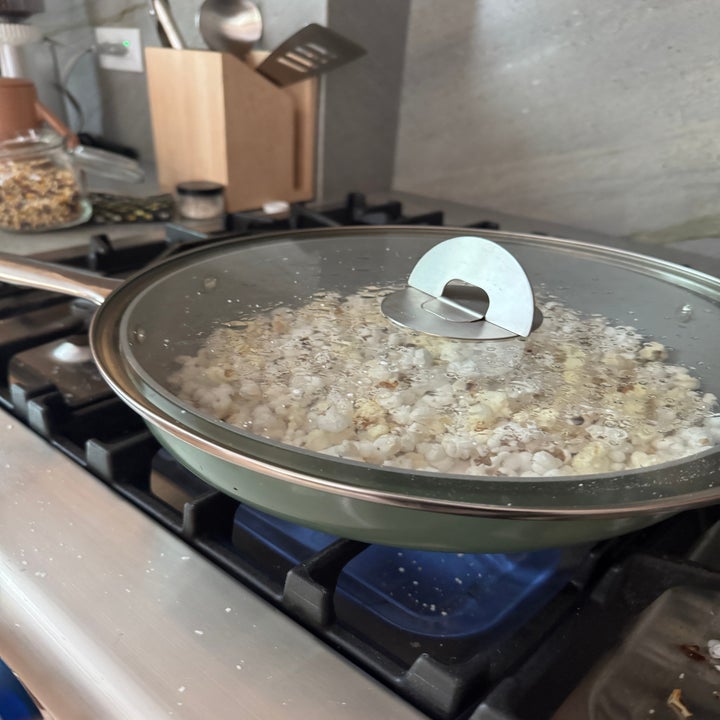 Popcorn popping in a covered pan on a stove, with kernels visible through the lid. Kitchen utensils are in the background