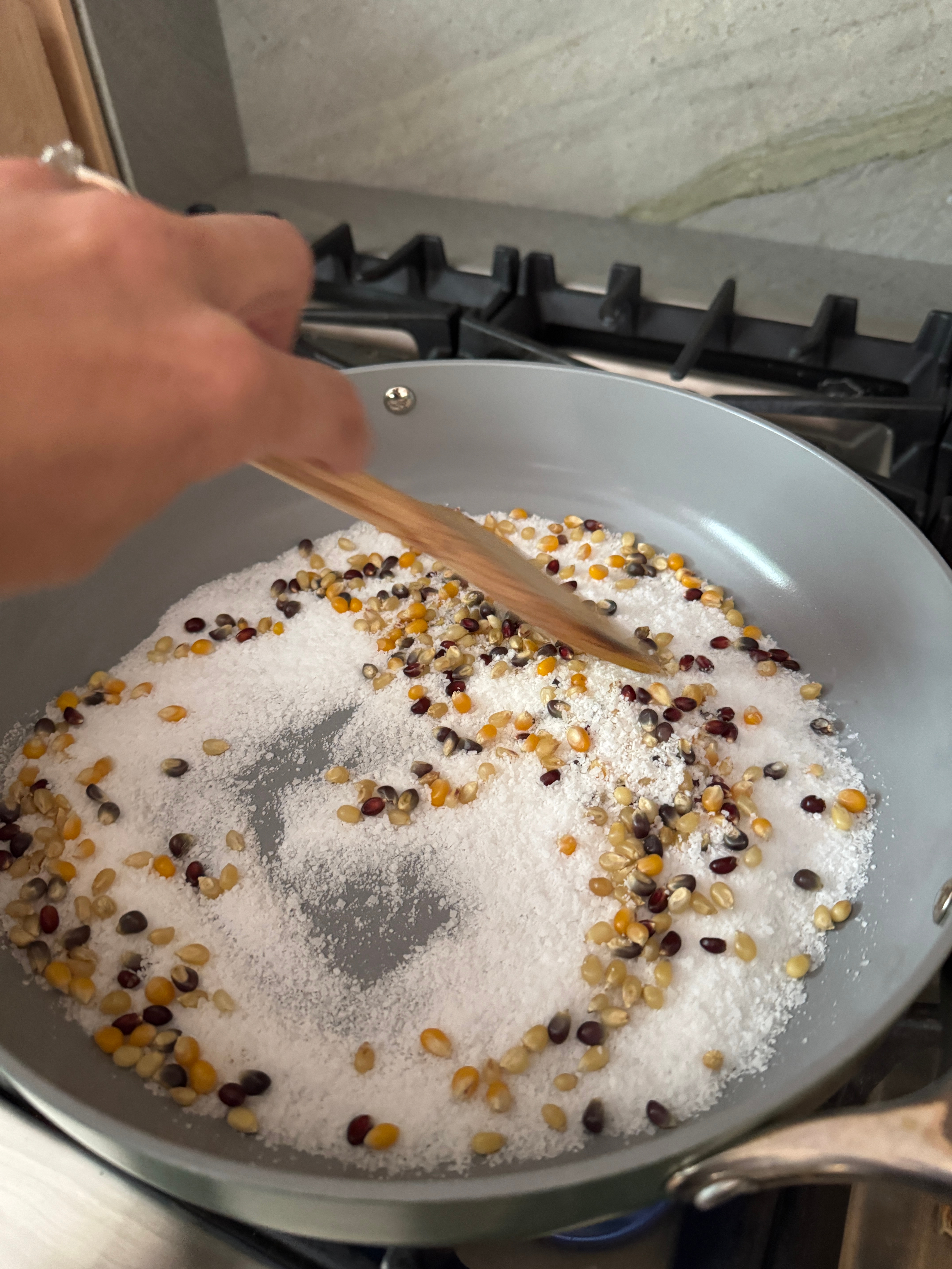 Hand stirring a mix of colorful popcorn kernels and sugar in a pan on a stove