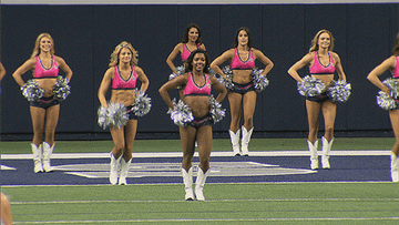 Cheerleaders perform a routine on a sports field, wearing crop tops, shorts, and white boots, while holding silver pom-poms