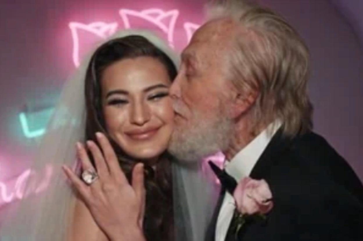 A bride smiles as an older man in a tuxedo kisses her cheek in front of neon lights