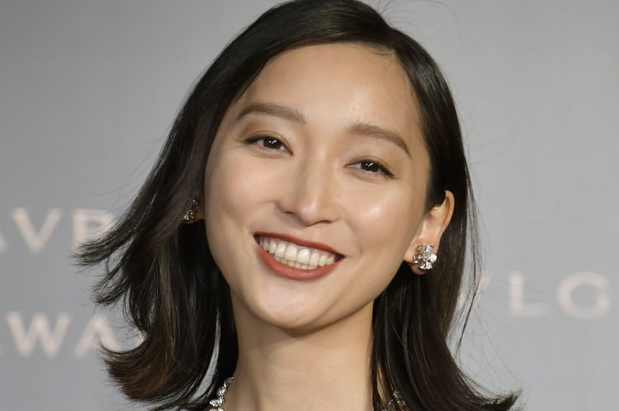 有名人がフォーマルなドレスを着て微笑んでいる。宝石のネックレスとイヤリングを身に着け、シンプルな背景の中で。