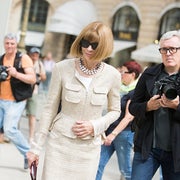 Person in a tailored, textured jacket and skirt set, wearing sunglasses and a necklace, walking past photographers