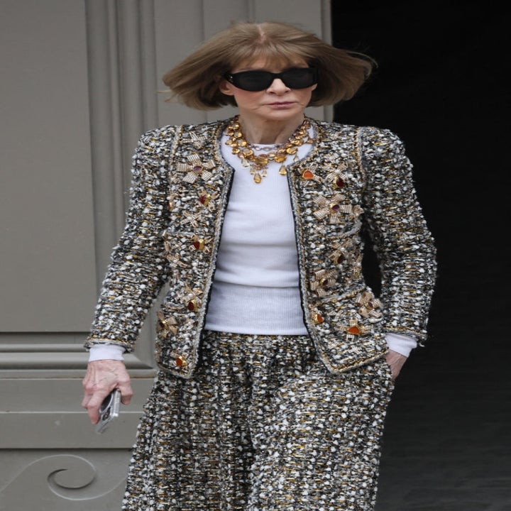 Person wearing ornate patterned outfit, large sunglasses, and statement necklace, holding a phone while walking outdoors