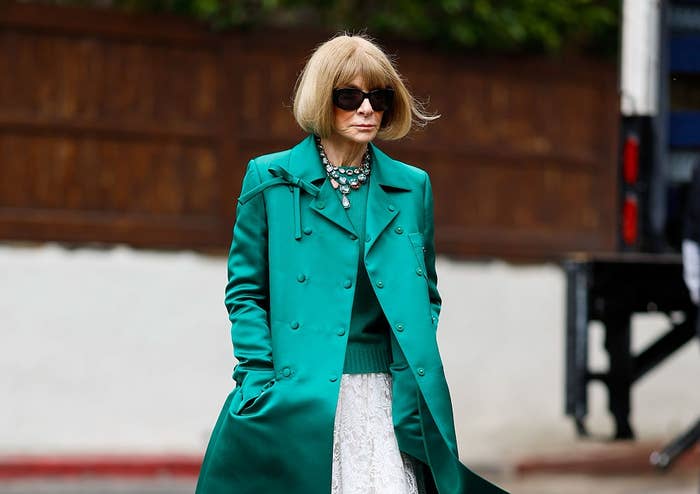 A person in a stylish trench coat, sunglasses, and layered necklaces is walking outside with hands in pockets, exuding a chic, polished vibe