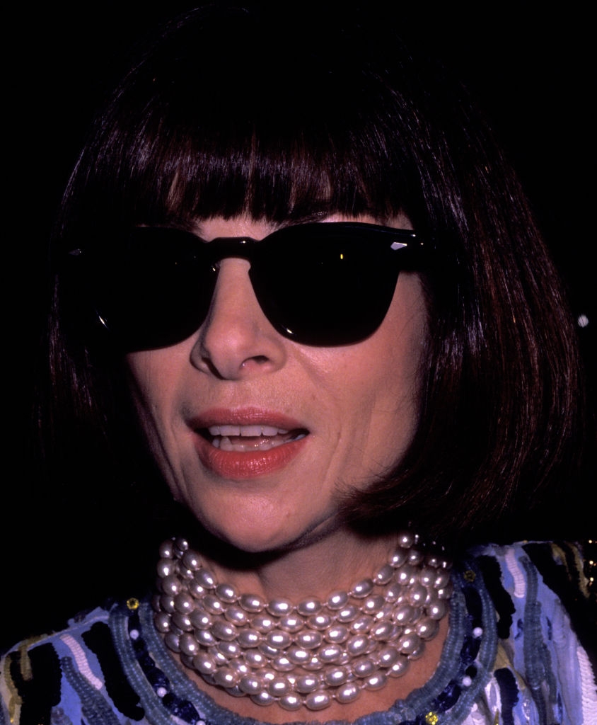 Person wearing dark sunglasses, a short bob haircut, and a multi-layered pearl necklace, dressed in patterned attire