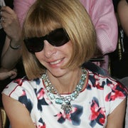 Person with bob haircut wearing sunglasses and a floral-patterned dress with a jeweled neckline, smiling at an indoor event