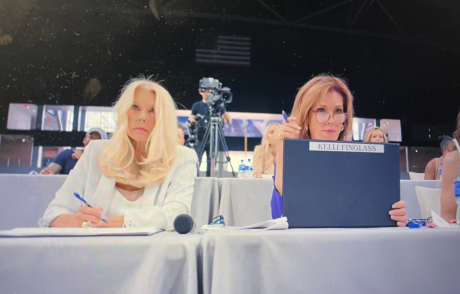 Two women are seated at a table with papers and a microphone, appearing focused. One holds a clipboard. A camera setup is visible in the background