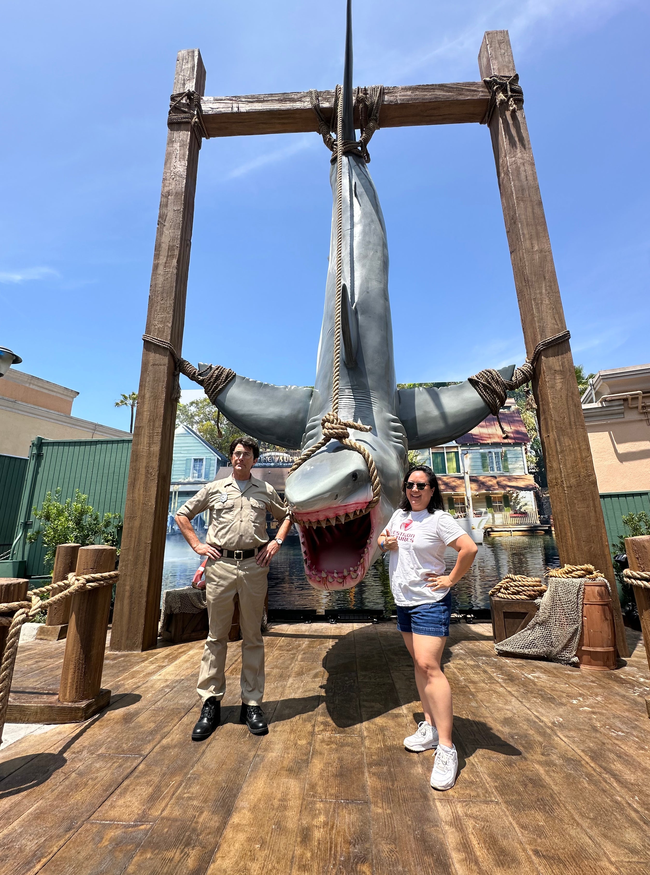 Person poses next to a large shark model displayed as an attraction, with a staff member dressed in a theme park uniform standing nearby