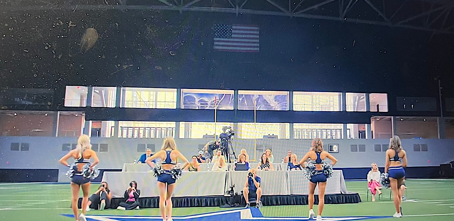 Cheerleaders in formation on an indoor field perform with judges seated at a table in the background. An American flag hangs above