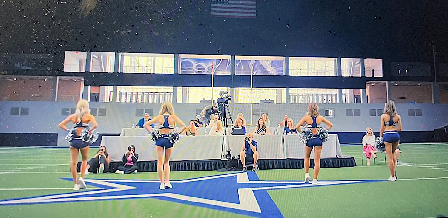 Cheerleaders in formation on an indoor field perform with judges seated at a table in the background. An American flag hangs above