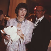 Woman in a sparkling suit smiles while holding papers; man in glasses stands nearby. Indoor event setting